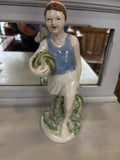 Porcelain figurine of a smiling boy holding a watermelon, wearing a blue vest and white shorts, standing barefoot on a leafy green base.