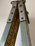 Close-up of vintage folding measuring stick with metal hinge and bold black and red markings on yellow background.