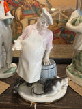 Porcelain figurine titled “The Cook and the Cat” by Lomonosov, depicting a weary cook in apron and chef’s hat beside a barrel, with a cat at his feet. A colourful vintage print is in the background.