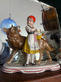 Porcelain figurine of Little Red Riding Hood standing beside the Wolf, with bright hand-painted details including a red headscarf, yellow basket, and a decorative base with red scrollwork.