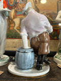 Rear view of the Lomonosov figurine showing the cook leaning heavily on a blue barrel, wearing brown trousers and black boots, with the cat partially visible behind.