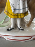 Detail of the girl’s lower half and the base of the figurine, highlighting her white stockings, black shoes, and yellow-trimmed dress with hand-painted embellishments.