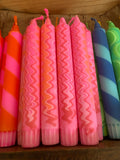 Close-up of vibrant pink candles with bold orange and red zigzag patterns, neatly arranged in a row with glimpses of striped and swirled candles in various colours on either side.