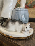 Close-up of the Lomonosov porcelain cat figure crouching beside the cook, shown nibbling on a scrap of food, with detailed hand-painted shading and glossy finish.