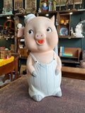 Front view of mid-century bisque pig money bank showing full figure in dungarees with painted facial features and flat cap.