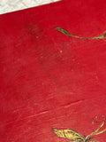 Close-up detail of the red painted tabletop surface showing light surface wear and scratching consistent with age, with partial gilt botanical stem and leaf motif visible at the edges.