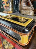Angled close-up of Victorian japanned cash box showing black lacquered lid with mustard yellow frame, brass twin knob handles and rolled tan banding around body