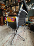 Eames EA108 chair viewed from behind at an angle, showing chrome frame detail and black fabric.
