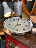 Front view of 1970s Treasure oval alarm clock in chrome case showing cream dial with bold black Arabic numerals, luminous green-tipped hands, red seconds hand and red Treasure brand logo, displayed on wooden shelf