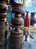 A close-up of a dark brown carved wooden decorative object with stacked circular tiers and textured bands, displayed on a reflective surface with other colorful vases in the background.