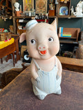 Vintage bisque porcelain pig money bank in blue-striped dungarees and flat cap with hand-painted eyes and open mouth, three-quarter view.