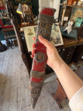 Hand holding the upper scabbard, showing the suspension rings, figural relief panel, and red painted body leading down to the portrait medallion.