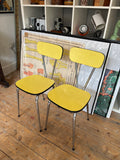 Pair of vintage mid-century yellow Formica dining chairs with chrome hairpin legs, side view against a shelving unit.