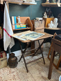 The antique folding table styled in an antique shop setting with Russian posters laid on top, surrounded by books, ceramics and curiosities, showing the table in use at a comfortable working height.