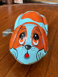 Vintage tin wind-up jumping dog toy, front view showing big-eyed puppy face in orange and pale blue.