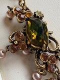 Macro detail of a single Florenza necklace link showing a large faceted peridot green marquise rhinestone in an ornate antiqued gold scrollwork setting with amber rhinestones and faux pearl drops.