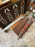 The sword drawn from its scabbard, both laid out together on a wooden bench, showing the full etched blade and matching red and metal scabbard.