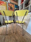 Pair of vintage yellow Formica dining chairs viewed from behind, showing chrome crisscross back supports.