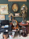 Wide shot of an antique shop interior showing the large oval Queen Victoria portrait hanging prominently on the wall above a varied display of antiques including furniture, mirrors, and decorative objects.