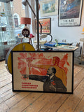 Framed 1969 Soviet propaganda poster by V. Konuhov leaning against the floor in a vintage shop, showing Lenin pointing forward and a Soviet worker holding a flaming torch aloft, with Cyrillic text reading "Победа коммунизма неизбежна!" (The Victory of Communism is Inevitable). Black frame, horizontal format.