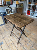 Antique Victorian folding table with turned notched legs, three-quarter front view showing worn planked top with rounded corners, dark stained X-frame scissor base with double stretcher rails, standing open on bare wood floor.