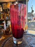 Large Dartington Crystal cranberry glass vase, full height view, showing deep ruby-red optical ribbed body with clear crystal base, displayed in an antique shop window setting.