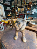 Front-on view of the vintage ceramic tiger planter in antique shop setting, showing the full face with hand-painted eyes and whiskers, mustard yellow saddle planter visible behind the head, and sturdy modelled front legs.