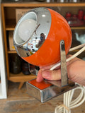 Hand holding vintage orange 1970s space age eyeball desk lamp showing chrome reflector and tilting head