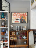 The framed Konuhov poster hanging on a grey wall in a vintage shop, displayed above a mid-century oak display cabinet. Surrounding items include a large wall clock, vintage ceramics on shelves, chrome canisters and a small mantle clock on top of the cabinet.