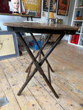 Side view of the antique folding table showing the X-frame scissor base fully open, turned notched legs crossing at centre, and the dark stained underside of the planked top with original wooden catch visible.