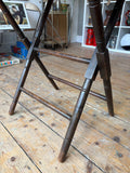Close-up of the base of the antique folding table showing the dark stained turned legs with notched ring detailing, double horizontal stretcher rails, and the lower crossing point of the X-frame mechanism.