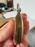 A close-up of a hand holding a vintage pocket watch, showing its side profile with a silver-toned metal case and a gold-toned center plate.