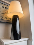 Habitat Joséphine table lamp lit showing warm cream glow through white glass shade with black and white glass base visible below, displayed in shop corner