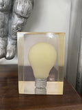 Clear amber resin block containing a suspended frosted incandescent lightbulb with metal screw base, front view.