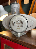 Rear view of Treasure oval alarm clock showing grey metal back plate with alarm, time-set and wind controls, battery compartment and splayed chrome foot stand