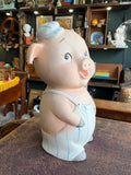 Left side view of bisque porcelain pig bank showing profile, ear, flat cap and dungaree straps.