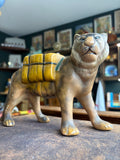 Vintage mid 20th century ceramic tiger planter, three-quarter front view showing standing tiger in matte sandy grey-brown glaze with hand-painted facial features and open mustard yellow saddle planter on its back with teal blue strap detail.