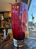 Dartington Crystal cranberry vase seen from a slightly lower angle, emphasising the cylindrical form and rich colour depth with shop interior reflected through the glass.