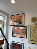 The framed Konuhov Soviet propaganda poster hanging on a white wall alongside two other framed vintage posters — a Chinese Cultural Revolution poster below it and a black-and-white Chinese newspaper broadsheet to the right. Photographed from a low angle near a staircase.