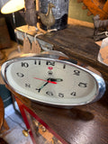 Slightly elevated front view of vintage Treasure oval chrome alarm clock showing full dial with bold numerals, luminous hands and red alarm hand, on dark wooden surface