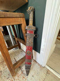 Full-length view of a vintage cast metal Roman gladiator sword prop in its scabbard, leaning against a wooden stool, showing lion-head pommel and red painted scabbard body.