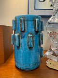Turquoise crackle glaze cylinder vase with raised rectangular blocks and brown highlights, on a wooden sideboard