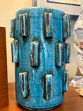 Turquoise pottery vase with brutalist raised block decoration and dark brown highlights on a teak surface